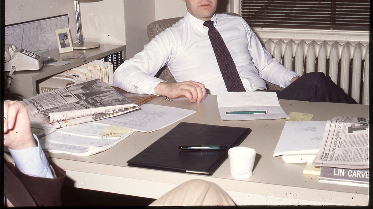 Man in tie sits at desk covered with papers/documents, another man sits mostly off camera to left